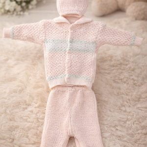May include: A light pink knitted baby outfit, including a jacket, pants, and a hat. The jacket has a collar and a light blue stripe across the chest. The outfit is displayed on a fluffy, white surface.