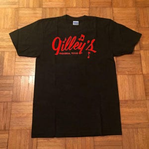 Może przedstawiać: Czarny t-shirt z krótkim rękawem z czerwonym napisem Gilley's oraz Pasadena, Texas nadrukowanym na klatce piersiowej. Wzór zawiera małe czerwone ikony nut muzycznych wkomponowane w liternictwo. Koszulka ma standardowy okrągły dekolt.