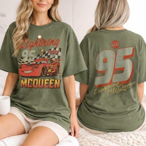 Puede incluir: Camiseta verde oliva con un dise&ntilde;o de Rayo McQueen de la pel&iacute;cula Cars. El frente muestra "Lightning 95 McQueen" con una bandera a cuadros. La parte trasera tiene un gran "95" y "Lightning McQueen".
