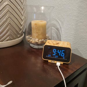 May include: A wooden digital alarm clock displaying the time 9:46 in blue digits. The clock is plugged in with a white cord. A candle in a glass vase and a decorative pot are in the background.
