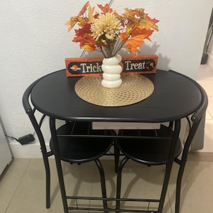 May include: A black round dining table with two matching chairs. The table is set with a decorative arrangement of fall leaves and flowers in a white vase, a woven placemat, and a small sign that says "Trick Treat".