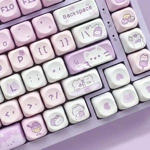 May include: A pastel purple mechanical keyboard with white and light purple keycaps. The keycaps feature a mix of standard letters and numbers, along with custom illustrated keys with cat and dessert designs. The keyboard has a silver volume knob.