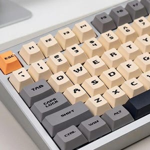 May include: A mechanical keyboard with a silver frame and beige and gray keycaps. The keyboard has an orange ESC key and a dark blue spacebar. Several loose keycaps are scattered around the keyboard.