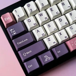 May include: A black mechanical keyboard with white and purple keycaps. The keycaps have white lettering and symbols, with some keys in Korean. The Esc key is pink, and the space bar has a pink and white design.