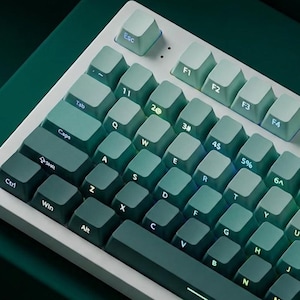 Pode incluir: Um teclado mecânico com estrutura branca e teclas gradientes em tons de verde-azulado. As teclas apresentam letras e números brancos. O teclado tem um layout compacto com teclas de função e setas.