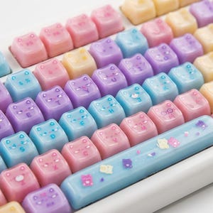 Puede incluir: Un teclado blanco con teclas personalizadas en colores pastel. Las teclas tienen forma de gato y presentan letras y símbolos. La barra espaciadora es azul claro con decoraciones de gatos y estrellas.
