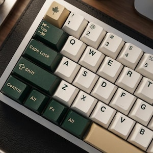 Puede incluir: Un teclado mecánico con marco plateado y una combinación de teclas blancas, verdes y beige. Las teclas presentan letras, números y símbolos estándar. Las teclas verdes tienen letras blancas y las beige un símbolo de hoja de arce.