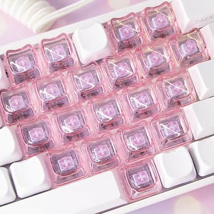 Pode incluir: Close-up de um teclado mecânico branco com teclas rosa transparentes. As teclas quadradas têm um símbolo de cruz no centro. Um cabo branco enrolado é visível.