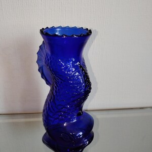 May include: A deep blue glass vase shaped like a fish. The vase features a textured surface, a flared rim with a scalloped edge, and a rounded base. The fish's tail and fins are sculpted on the side of the vase.