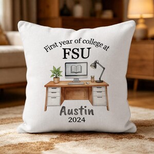 Op de afbeelding: Een wit vierkant sierkussen met een gedrukte illustratie van een houten bureau met een computer, een plant in een pot en een lamp. De tekst luidt First year of college at FSU, Austin, en 2024. Het ontwerp is gecentreerd op de voorzijde.