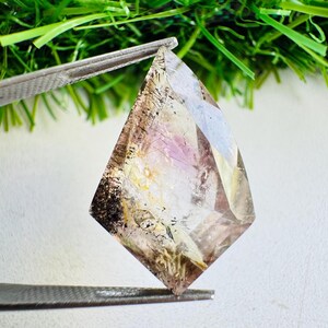 以下が含まれることがあります： 黒、金、紫の鉱物沈殿物の内部インクルージョンを特徴とする、凧形のファセットカットされた透明なクォーツの宝石。石は金属製のピンセットで保持されており、その透明な構造の中に複雑で多次元的なパターンを表示しています。