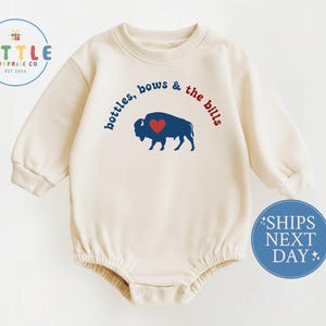May include: Cream-colored baby bodysuit with long sleeves. The front features a blue buffalo with a red heart and the text "bottles, bows & the bills" in blue and red. The bodysuit has a round neckline and snap closures.