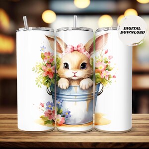 May include: White tumbler featuring a watercolor illustration of a bunny wearing a pink bow, peeking out of a blue bucket filled with flowers. The tumbler has a straw and a lid. The words "DIGITAL DOWNLOAD" are on the image.
