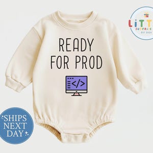 May include: A cream-colored long-sleeve baby bodysuit featuring the black text READY FOR PROD above a graphic of a purple computer monitor displaying code brackets. The garment has a crew neckline and ribbed cuffs at the wrists and leg openings.