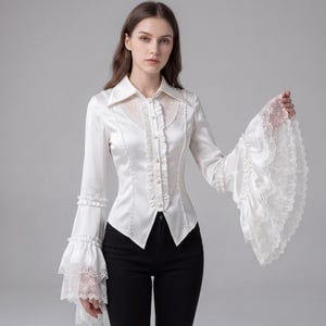 May include: A white satin blouse with long sleeves and a button-down front. The blouse has lace detailing on the collar, cuffs, and sleeves. The sleeves have a flared, bell-like shape. The blouse is worn with black trousers.