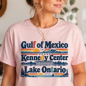 May include: A light pink short-sleeved t-shirt featuring a graphic print with the text Gulf of Mexico, Kennedy Center, and Lake Ontario. The design includes colorful horizontal stripes, a sun, a wave, a building, trees, and a sailboat.