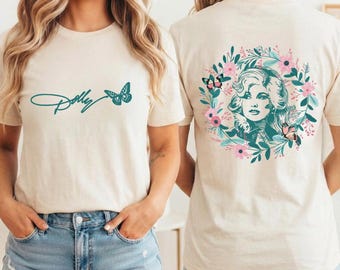 May include: A cream-colored short-sleeve t-shirt featuring a teal script logo reading Dolly with a butterfly on the front. The back displays a circular floral wreath graphic surrounding a portrait of a woman with butterflies in pink and teal.