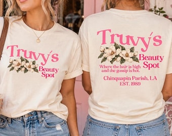 May include: A cream-colored t-shirt featuring pink text that reads Truvy's Beauty Spot with a magnolia flower graphic. The back of the shirt includes the additional text: Where the hair is high and the gossip is hot. Chinquapin Parish, LA EST. 1989.