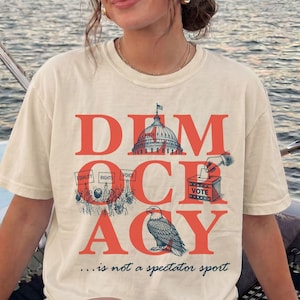 Op de afbeelding: Een ivoorkleurig T-shirt met korte mouwen en een grote rode afbeelding met het woord DEMOCRACY. Het ontwerp bevat een illustratie van het Capitool, een stembus, een menigte met borden en een Amerikaanse zeearend. De tekst luidt ...is not a spectator sport.