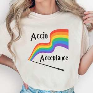 May include: Ivory-colored t-shirt featuring a rainbow flag design with the words "Accio Acceptance" and a wand. The design is in the center of the shirt. The shirt is paired with blue denim shorts.
