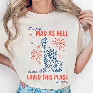 Op de afbeelding: Een ivoorkleurig T-shirt met korte mouwen en een blauwe afbeelding van het Vrijheidsbeeld, omringd door rood vuurwerk. De tekst luidt: I'm just MAD AS HELL cause I LOVED THIS PLACE Est. 1776. Het T-shirt wordt gedragen met een lichtblauwe spijkerbroek.
