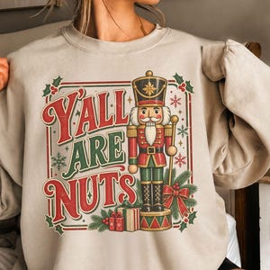 May include: A sand-coloured crewneck sweatshirt featuring a graphic print of a nutcracker soldier standing on a base with gifts. The text reads Y'ALL ARE NUTS in red and green letters, framed by holly leaves, berries, and snowflakes in a rectangular border.