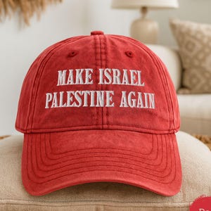 Puede incluir: Gorra de béisbol roja con el texto bordado "MAKE ISRAEL PALESTINE AGAIN" en blanco. La gorra está sobre un cojín beige. La gorra tiene una visera curva y un diseño de seis paneles.