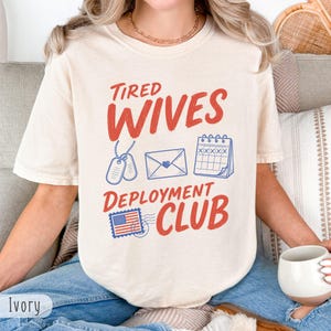 Könnte beinhalten: Elfenbeinfarbenes T-Shirt mit dem roten Text "Tired Wives Deployment Club". Das Shirt zeigt Illustrationen von Erkennungsmarken, einem Umschlag, einem Kalender und einer Briefmarke.