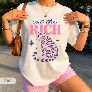 May include: An ivory t-shirt featuring a graphic print of a tiger with purple stripes. Above the tiger, the text reads "eat the" in dark blue and "RICH" in large pink letters. The design includes small four-pointed star accents in dark blue.