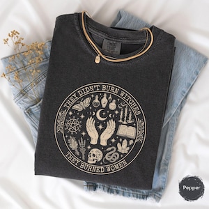 May include: Dark gray t-shirt with the text "THEY DIDN'T BURN WITCHES THEY BURNED WOMEN" in a circular design. The design includes hands, a skull, a snake, and other mystical symbols. A gold necklace rests on the shirt, which is folded on top of blue jeans.