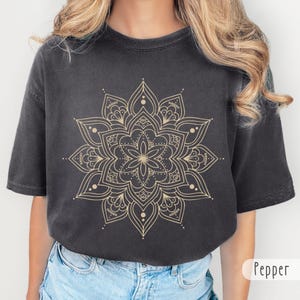 May include: A charcoal gray short-sleeve t-shirt featuring a large, intricate gold-colored mandala graphic printed on the chest. The symmetrical design includes repeating floral-like patterns and the repeated text "RESIST" integrated into the artwork.