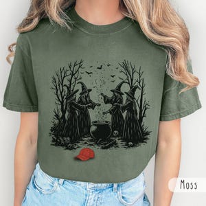May include: A moss green t-shirt featuring a black graphic print of four witches in pointed hats standing around a bubbling cauldron in a forest setting with bats overhead. A small, bright red baseball cap is illustrated at the bottom center.