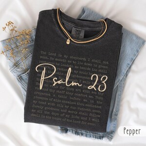 May include: A charcoal grey crewneck t-shirt featuring the full text of Psalm 23 printed in a small, dark font, with the words "Psalm 23" overlaid in a large, cream-coloured script font. The shirt has a vintage-style wash and a Comfort Colors label.