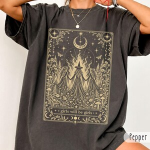 Könnte beinhalten: Ein anthrazitfarbenes Kurzarm-T-Shirt mit einem rechteckigen beigen Grafikdruck. Das Design zeigt drei Figuren in langen Kleidern und Hexenhut in einem Wald mit Pilzen, Sternen und einer Mondsichel sowie dem Schriftzug girls will be girls.