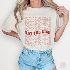 Peut inclure: T-shirt de couleur ivoire avec l'inscription "EAT THE RICH" dans une police rétro répétée. Le texte est principalement rouge, avec une ligne dans une nuance plus foncée. Le t-shirt est porté avec un short en jean.