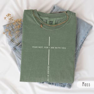Puede incluir: Una camiseta de manga corta color verde musgo con un gráfico de cruz vertical blanca. El texto dice FEAR NOT, FOR I AM WITH YOU e ISAIAH 41:10. La camiseta tiene un cuello redondo acanalado con una etiqueta Comfort Colors en el interior.