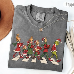 Könnte beinhalten: Ein anthrazitfarbenes Kurzarm-T-Shirt mit einem Grafikdruck einer Gruppe von Figuren in rot-grüner Festtagskleidung, die über einen Zebrastreifen gehen, wobei eine grüne Figur hinterherläuft. Das Shirt hat einen Rundhalsausschnitt und ein Comfort Colors-Etikett.
