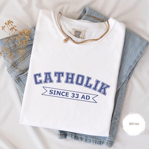 May include: A white t-shirt featuring the word "CATHOLIK" in blue, above a banner reading "SINCE 33 AD". The shirt is folded on top of blue denim jeans. A gold necklace and a Comfort Colors tag are also visible.