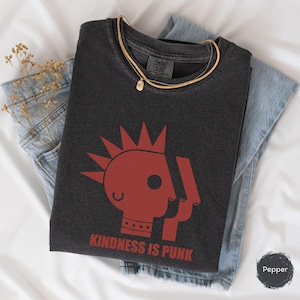 Puede incluir: Camiseta gris oscuro con un gráfico rojo de una cara estilizada con cresta y el texto "KINDNESS IS PUNK". La camiseta está doblada sobre unos vaqueros azules, con un collar dorado y flores secas.