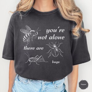 Puede incluir: Camiseta gris oscuro con ilustraciones blancas de una abeja, un escarabajo y un grillo. El texto "you're not alone there are bugs" está impreso encima y debajo de los dibujos de insectos. La camiseta es de estilo casual.