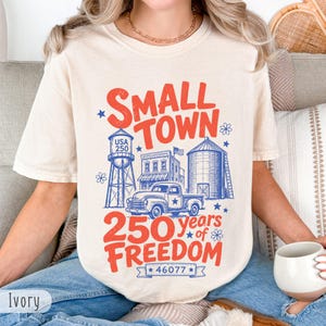 Op de afbeelding: Ivoor kleurig t-shirt met een rood en blauw ontwerp. De afbeelding toont "Small Town" boven een illustratie van een watertoren, gebouwen en een vrachtwagen. Daaronder staat "250 years of Freedom" en het nummer "46077".