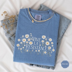 May include: A blue jean-colored t-shirt with the phrase "Vive La Resistance" in white script, surrounded by a floral daisy design. The shirt is folded with a gold necklace and a pair of blue jeans.