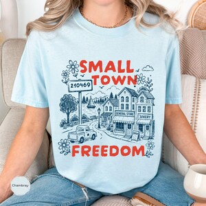 May include: A light blue t-shirt featuring a dark blue line art illustration of a small town scene with a truck, buildings, and a sailboat. The text "SMALL TOWN" and "FREEDOM" is printed in bold red letters, and a sign displays the number 210469.