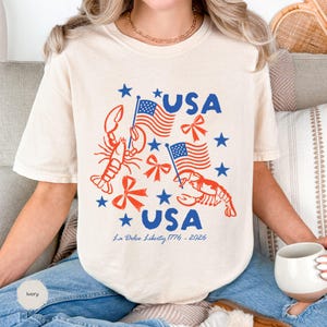 Puede incluir: Camiseta de color marfil con un diseño patriótico. Presenta la palabra "USA" en azul, banderas americanas, langostas rojas y estrellas azules. El texto "La Dolce Liberty 1776-2026" está impreso debajo.