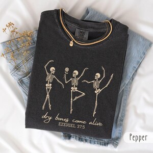 Peut inclure: Un t-shirt gris anthracite avec un motif de trois squelettes dansants, dont l'un tient une fleur. Le texte "dry bones come alive" et "EZEKIEL 37:5" est imprimé en beige clair. Le t-shirt présente un col rond et une étiquette Comfort Colors.