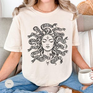 May include: Ivory-colored t-shirt featuring a Medusa-inspired graphic. The design includes a woman's face surrounded by snakes, with words like "Freedom," "Courage," and "Wisdom" encircling the image. The shirt is made of a soft material.