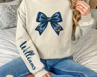 May include: Sand-colored sweatshirt with a navy blue bow design, an anchor, and the text "MOM EST. 2026." The left sleeve features the name "Williams" in navy blue. The sweatshirt is a casual style.