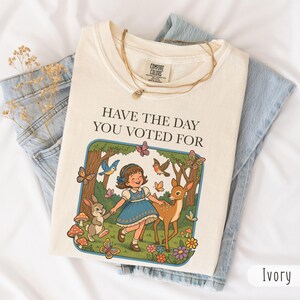 Puede incluir: Sudadera Comfort Colors color marfil con el texto "Have the Day You Voted For" y una ilustración caprichosa de una niña, un ciervo, un conejo, mariposas y pájaros. La sudadera está doblada sobre unos vaqueros azules.