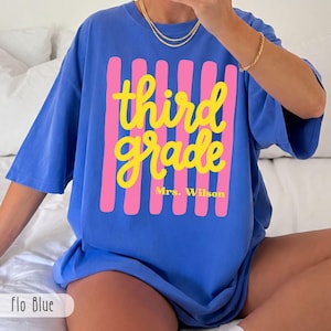 Op de afbeelding: Een blauw t-shirt met een afbeelding van verticale roze strepen, de gele tekst "third grade" in een cursief lettertype en "Mrs. Wilson" in een kleiner schreefloos lettertype eronder. Het shirt heeft korte mouwen en een ronde hals.