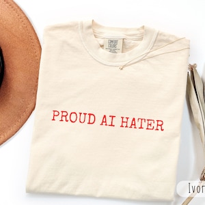 Może przedstawiać: T-shirt z krótkim rękawem w kolorze kości słoniowej z napisem PROUD AI HATER wydrukowanym czerwonymi wielkimi literami na klatce piersiowej. Koszulka ma okrągły dekolt i metkę marki Comfort Colors widoczną po wewnętrznej stronie kołnierza.